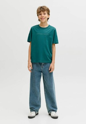 Niño de pie sonriendo, vestido con una camiseta verde de Jack & Jones, vaqueros azules holgados y zapatillas blancas sobre un fondo liso.