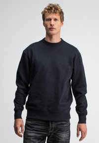 Marineblauwe sweatshirt met een gestructureerde afwerking, ronde hals en ribgebreide manchetten. Heeft een subtiel logo op de borst en een relaxte pasvorm.