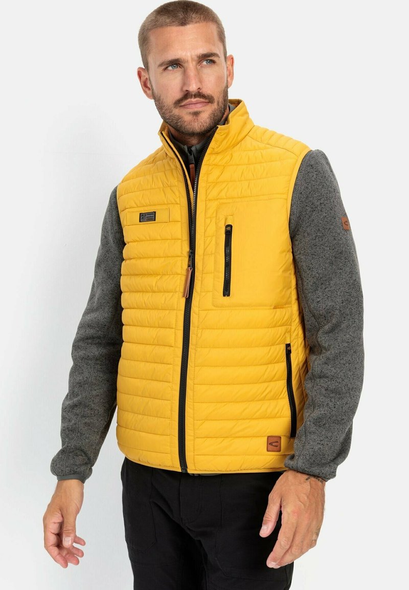 Quilted gul vest i gul med grå strikkede ærmer, med en lynlåslomme på brystet, sidelommer og et logo-patch på kanten.