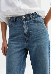 Niebieskie dżinsowe spodnie z wysokim stanem, zapinane na guzik, z pięcioma kieszeniami i kontrastującymi pomarańczowymi szwami. Gładka struktura i prosty krój.