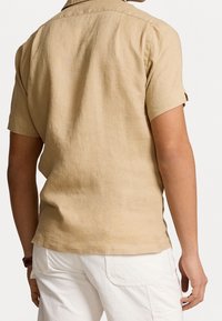 Camisa de lino beige de manga corta con un corte relajado, cuello abotonado y mangas enrolladas. Textura suave y diseño sencillo.