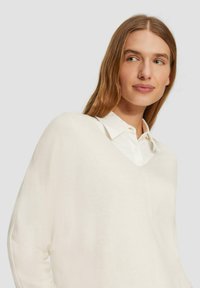 V-hals, lange mouwen trui in zacht, lichtgewicht crèmekleurig materiaal, gelaagd over een witte blouse met kraag. Minimalistisch ontwerp met een ontspannen pasvorm.