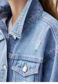 Světle modrá džínová bunda s velkým límcem, stříbrnými kovovými prvky, opotřebovanými detaily a dvěma náprsními kapsami. Měkká textura a klasický design.