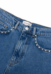 Μπλε τζιν από denim με ασημένιες τελείες στις μπροστινές τσέπες, πορτοκαλί ραφές και κούμπωμα με κουμπί πάνω από μια ετικέτα που γράφει OH APRIL.