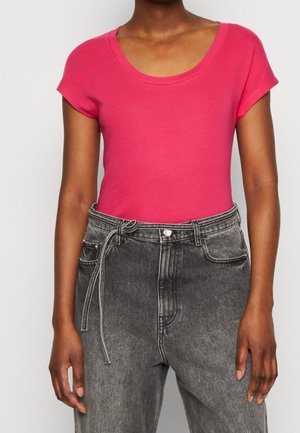 Woman wearing a bright pink short-sleeve top tucked into high-waisted gray faded jeans with a thin belt tied at the waist.