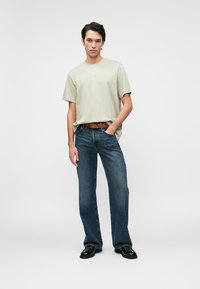 Light green cotton t-shirt with a relaxed fit, paired with dark blue denim flared jeans. Brown leather belt and black shoes. Plain background.