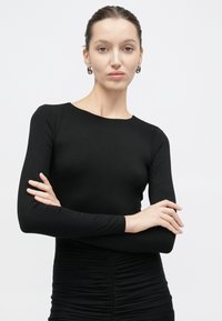 Γυναίκα με μαλλιά χτενισμένα πίσω, που φοράει ασημένια σκουλαρίκια κρίκους και εφαρμοστό μαύρο μακρυμάνικο τοπ με ρυτιδωμένη λεπτομέρεια, με σταυρωμένα τα χέρια.