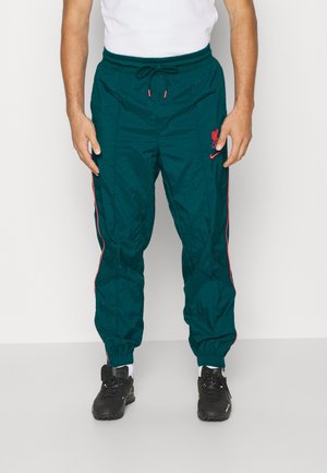 Mann trägt dunkelgrüne Trainingshose mit pinkfarbenem Besatz, Liverpool FC-Logo und schwarzen Sneakern, steht vor einem schlichten weißen Hintergrund.