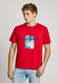 Red cotton t-shirt featuring a blue graphic print with "Pepe Jeans" text. Short sleeves and a round neckline. Casual fit with a smooth texture.