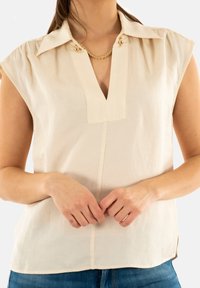 Blusa beige de manga corta con escote en V, que presenta pliegues en los hombros y botones decorativos en el cuello. Tejido suave y ligero.