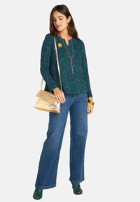 Grüne, floral gemusterte Bluse mit langen Ärmeln, blaue Jeans mit weitem Bein, grüne Schuhe, goldene Handtasche, goldener Schmuck, der das Outfit betont.