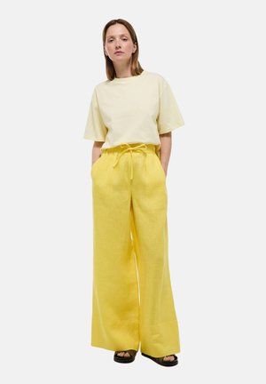 Femme debout, les mains dans les poches, portant un haut jaune pâle à manches courtes et un pantalon large jaune vif avec des sandales.