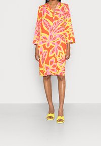 Buntes Blumenkleid mit orangefarbenen, rosa und gelben Mustern. Dreiviertel lange Ärmel, kragenloser Ausschnitt, knieumspielende Länge. Kombiniert mit gelben Sandalen mit Knoten.