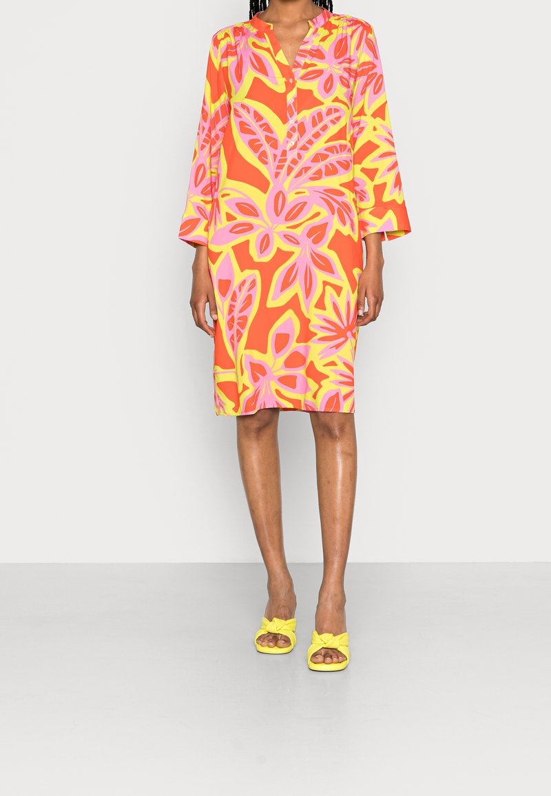 Buntes Blumenkleid mit orangefarbenen, rosa und gelben Mustern. Dreiviertel lange Ärmel, kragenloser Ausschnitt, knieumspielende Länge. Kombiniert mit gelben Sandalen mit Knoten.