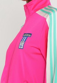 Chaqueta de chándal Adidas de un vibrante color rosa con rayas blancas y verde azulado en las mangas, cierre frontal con cremallera y parche con el logo de Adidas en el pecho.