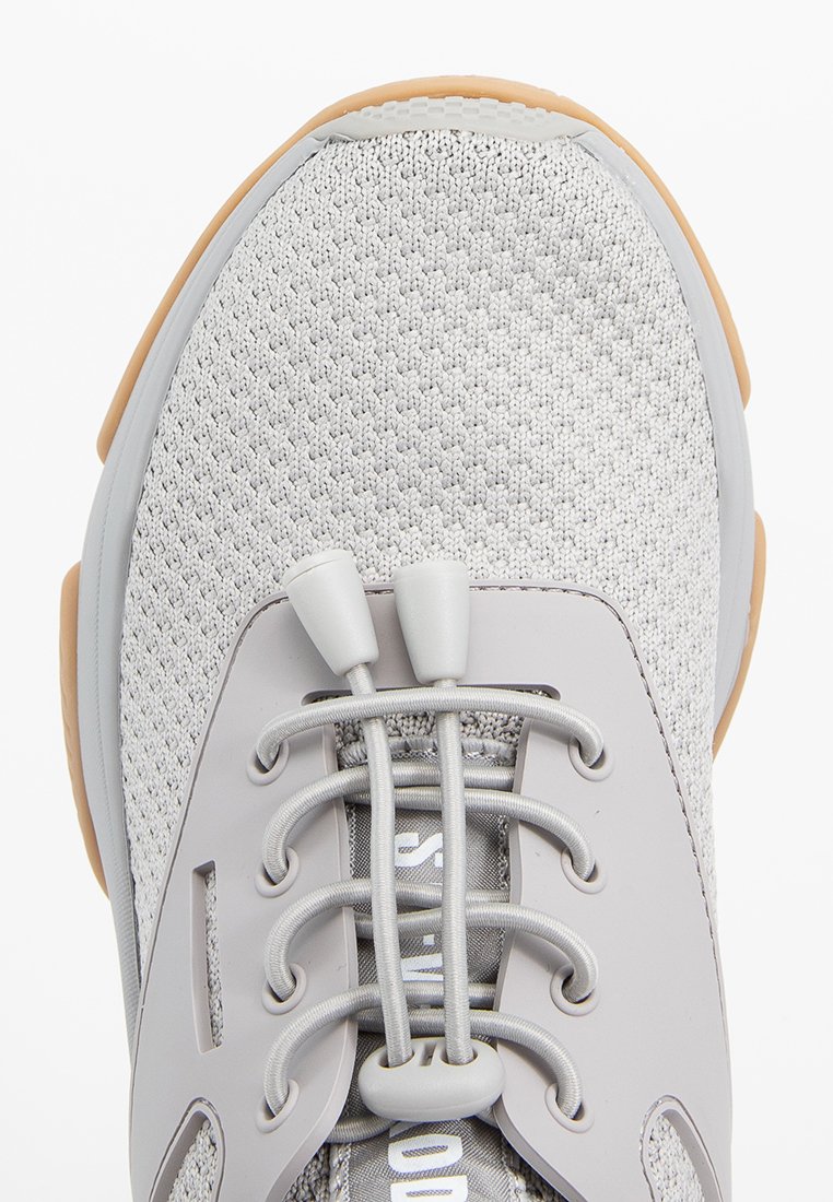 Primer plano de una zapatilla gris con parte superior de malla transpirable, cordones elásticos tipo bungee y borde de suela de goma en color marrón.