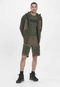 Mies, joka on pukeutunut oliivinvihreään ulkoilujakkuun, siihen sopiviin cargo-shorteihin, kuviolliseen t-paitaan ja mustiin vaelluskengiin, seisoo yksinkertaista taustaa vasten.