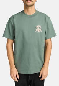 Green cotton t-shirt featuring a light pink graphic design of hands and sun on the left chest. Classic fit with a round neckline and short sleeves.