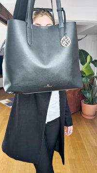Saco tote de couro preto com uma textura suave, apresentando duas alças longas e um detalhe circular dourado. Logotipo da marca na parte da frente.