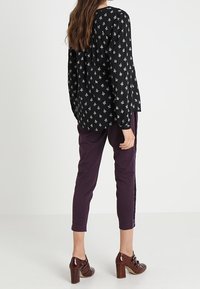 Blouse noire avec de petits motifs blancs, associée à un pantalon violet ajusté et des chaussures à talons carrés marron, vue de dos.