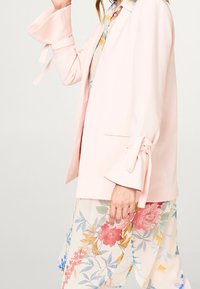 Kobieta w jasnoróżowym żakiecie z wiązanymi mankietami i spodniami w kwiatowy wzór w delikatnych pastelowych kolorach, stojąca na jednolitym tle.