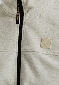 Veste matelassée gris clair avec fermeture éclair noire et un patch logo PME Legend près de la poitrine et du tirette de fermeture.