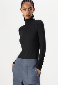 Schwarzes, geripptes Langarm-Turtleneck-Oberteil mit tailliertem Design, getragen mit hellgrauen, hochgeschnittenen Hosen, die ein Logo-Detail in der Nähe der Tasche haben.