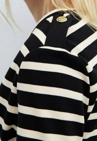 Maglione a righe orizzontali bianco e nero con una tracolla nera fissata da un bottone dorato, con parte dei capelli biondi in vista.