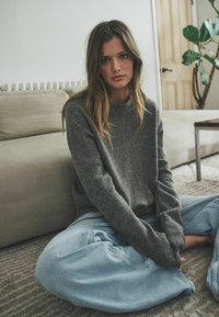 Sivý vlnený sveter s okrúhlym výstrihom a pohodlným strihom, zosynchronizovaný s svetlomodrými oversized riflemi, sediaci na textúrovanom koberci.