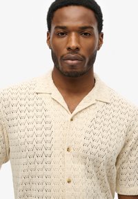 Beige kortærmet skjorte med knapper og et struktureret zigzag-mønster, lavet af letvægtsstof, med en camp krave og brune knapper.