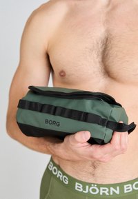 Green and black zippered toiletry bag with a matte finish, featuring a woven strap accent and a logo on the side. Rectangular shape.