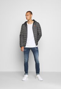 Giacca imbottita grigia con cappuccio, caratterizzata da cuciture orizzontali e un logo sul lato sinistro del petto, indossata sopra una maglietta bianca e jeans blu.