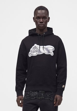 Homme portant un sweat à capuche noir avec un motif argenté de chat de dessin animé et un jean noir assorti avec des coutures texturées, debout devant un fond uni.