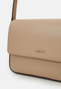 Béžová kabelka DKNY z texturované kůže, s klopou a viditelným stehováním podél okrajů. Na přední straně zlaté logo.