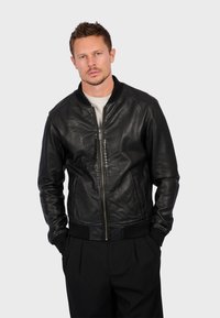 Černá kožená bomber bunda s ribovanými manžetami a spodním lemem; má přední zip a postranní kapsy. Hladká textura, klasický střih.