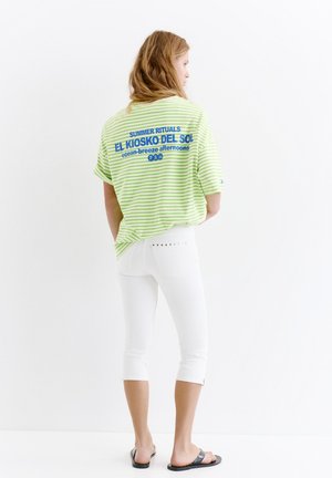 Femme aux longs cheveux portant un t-shirt rayé vert avec un texte bleu et un pantalon blanc court, debout devant un fond blanc.
