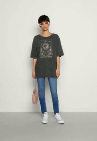 T-shirt cinza oversized com estampa gráfica, jeans azuis e ténis brancos. O modelo segura uma pequena bolsa rosa. Fundo simples.