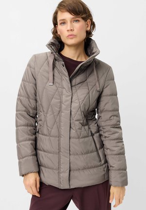 Woman wearing a taupe quilted jacket with a high collar, zip and snap buttons, standing against a plain light background.
