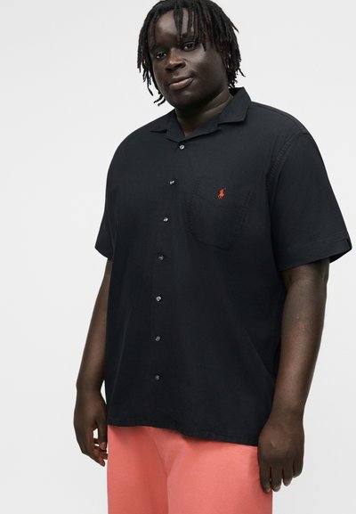 Mężczyzna z krótkimi dredami, ubrany w czarną, krótkorękawową koszulę na guziki z małym czerwonym logo i koralowe spodnie, stojący na jednolitym tle.