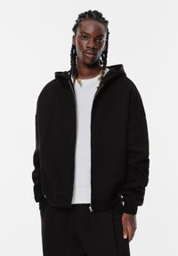 Felpa nera con zip sul davanti, cappuccio grande e vestibilità ampia. Indossata sopra un maglione bianco testurizzato, con spalle basse e tasche laterali.
