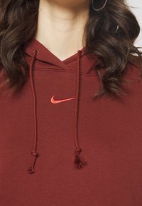 Mørkerød Nike-hoodie med snoretræk, rødbroderet logo, båret af en person med krøllet mørkt hår og sølv ørering i form af en ring.