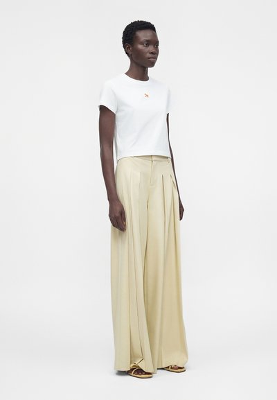 Γυναίκα όρθια, φορά λευκό τοπ με κοντά μανίκια και μικρό λογότυπο και μπεζ, πλισέ παντελόνι ψηλόμεσο με φαρδιά μπατζάκια και ανοιχτά σανδάλια.