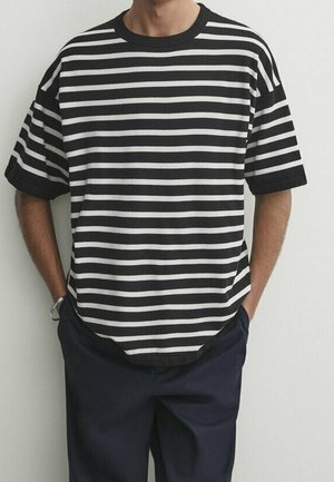 T-shirt à rayures noires et blanches avec des manches courtes, col rond, coupe décontractée, associé à un pantalon sombre. Le tissu semble lisse et léger.