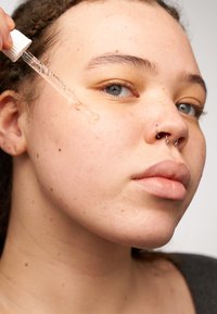 Klart dråpeflaske som dispenserer et halvtransparent serum på huden. Ansiktet har en jevn tekstur med fregner og har en subtil nesering.