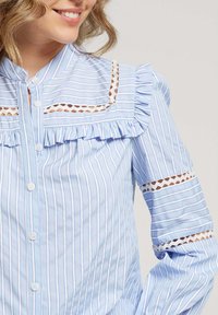 Chemise bleu clair à manches longues avec des rayures blanches verticales, un col froncé et des bords de manches, ainsi que des découpes en dentelle au niveau des épaules.