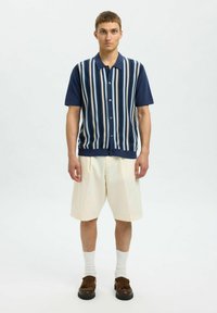 Prazna majica s mornarskim prugama kratkih rukava, s svijetloplavim i bež vertikalnim prugama, sa zatvaranjem na dugmad, u kombinaciji s krem kratkim hlačama i smešnim cipelama.