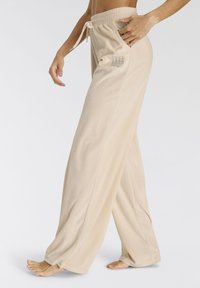 Beige weite Hosen mit einer glatten Textur, elastischem Bund, verstellbarem Kordelzug und einem dekorativen Logo auf der linken Seite.