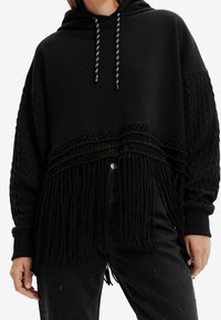 Sudadera negra con capucha que presenta mangas de punto texturizadas, un dobladillo trenzado y un remate con flecos. Cuerdas ajustables en la capucha.
