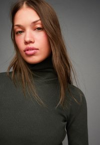 Dunkelgrüner gerippter Rollkragenpullover, Nahaufnahme, mit taillierter Passform und hohem Kragen. Langes, glattes Haar verdeckt teilweise den Ausschnitt.