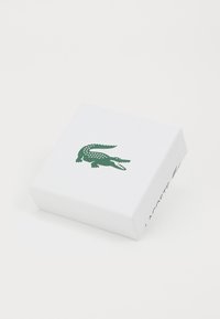 Boîte rectangulaire blanche avec une surface texturée, arborant un logo de crocodile vert sur le dessus et le mot "LACOSTE" imprimé sur le côté.